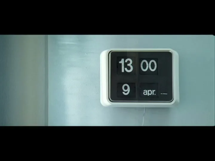 A white and black flip clock on a light blue wall shows the time changing from 12:59 to 13:00 on April 9th.