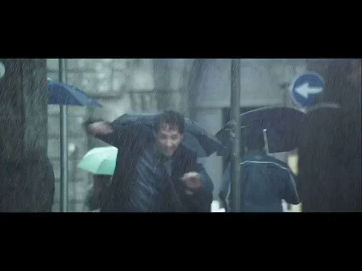 A man in a dark coat runs out of a building into a rainy city street, dodging people and a scooter, then almost gets hit by a car.