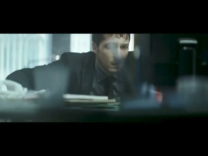 A man in a suit, with wet hair and a worried expression, leans over a desk in an office, looking intently at something out of frame.