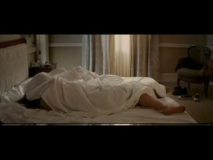 A person is seen lying in bed, covered by a white sheet, with one leg exposed, in a bedroom during the day.