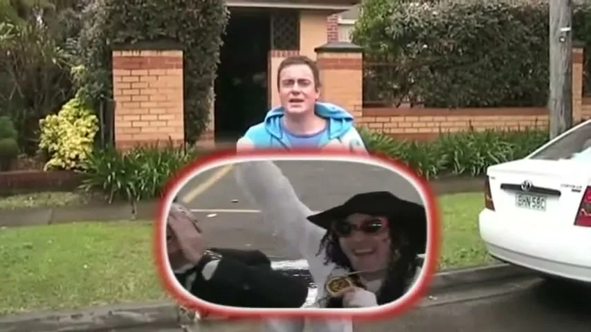 A young man in a blue hoodie stands on a suburban street, while a reflection in a car mirror shows two men in costumes.