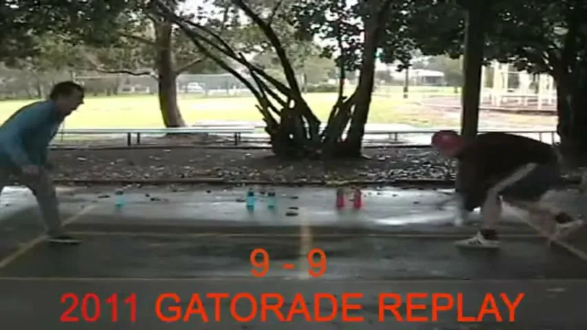 Two men play a game of throwing a block at bottles on a wet outdoor court, with a score of 9-9 displayed.