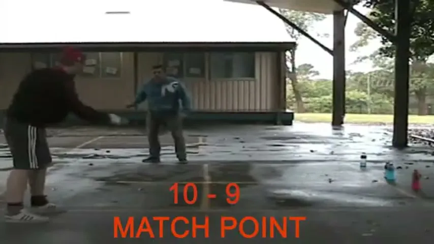 Two men play ping pong on a wet outdoor court, with a building and trees in the background, and a score overlayed on screen.