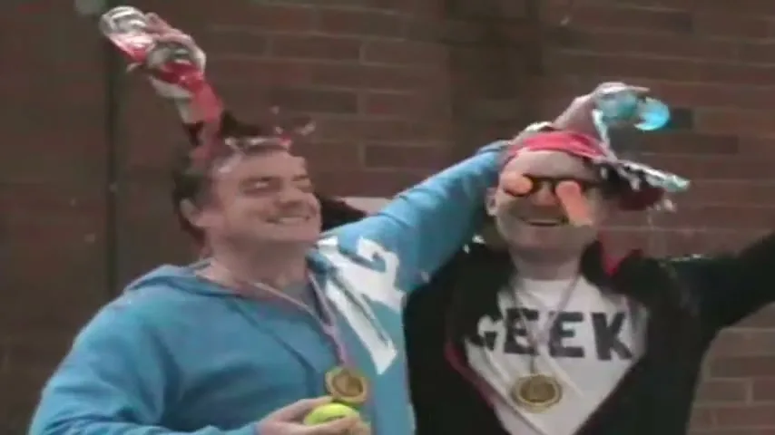 Two men, one in a blue hoodie and the other in a "GEEK" t-shirt, celebrate by pouring drinks on their heads and holding medals in front of a brick wall.