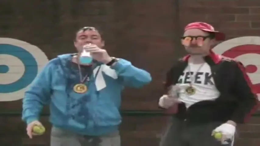 Two men, one in a blue hoodie and the other in a black jacket and red cap, celebrate with medals and Gatorade bottles in front of target signs.