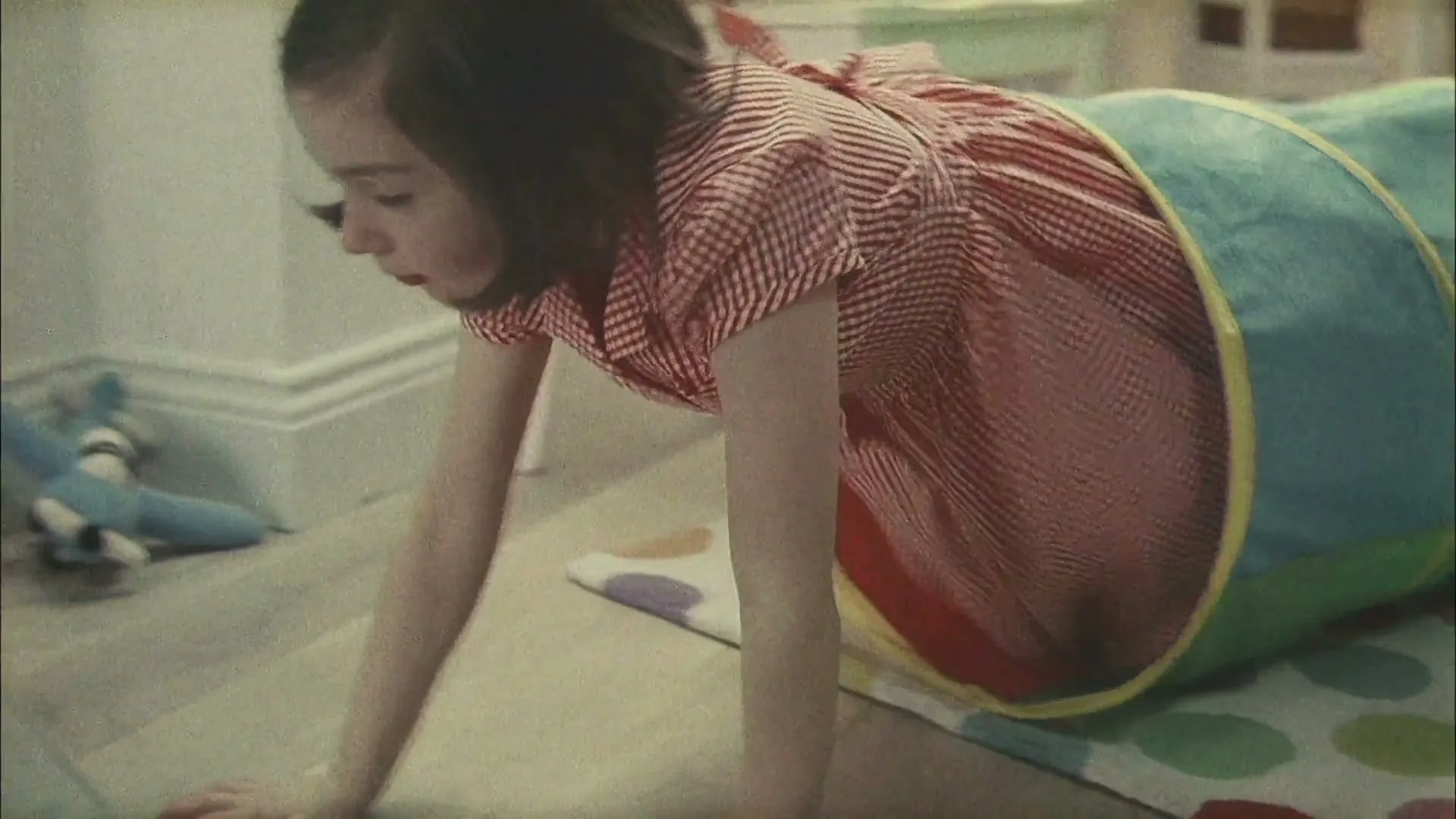 A baby is lifted from a crib, then a toddler crawls through a play tunnel, then a young girl sits at a school desk, then a girl at a birthday party.