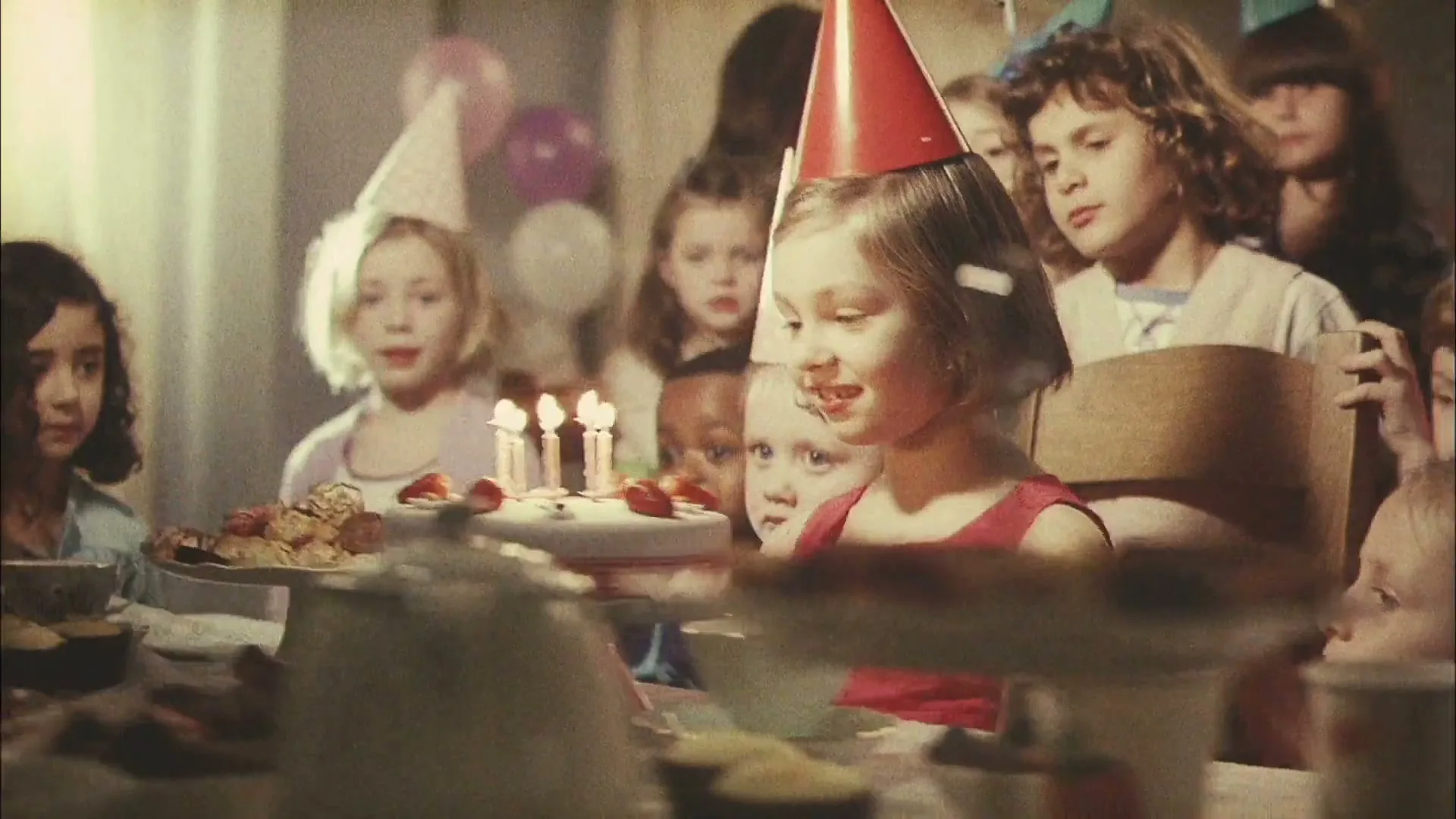A young girl in a red dress and party hat blows out candles on a birthday cake, surrounded by other children at a party table indoors.