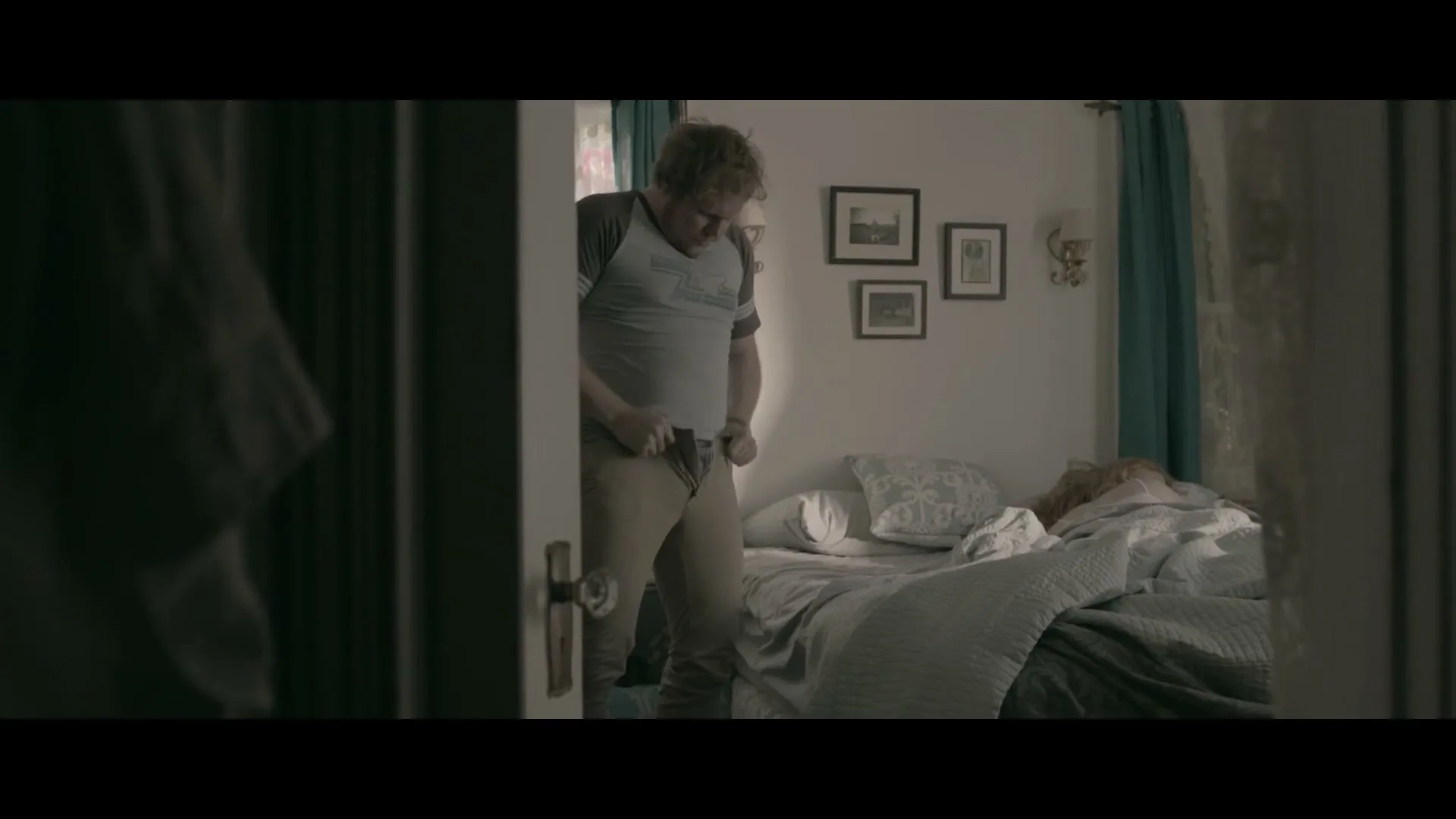 A man with messy hair and a t-shirt struggles to pull up his pants while a woman sleeps in bed in a bedroom.