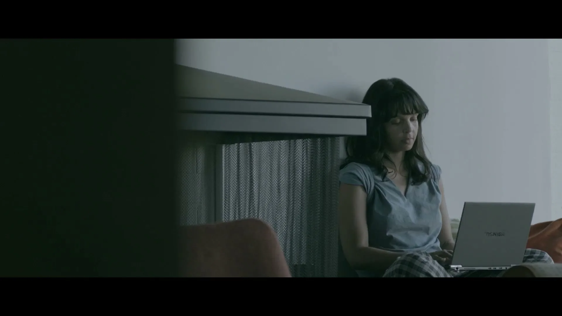 A young woman with dark hair sits on the floor, leaning against a wall, typing on a silver Toshiba laptop in a dimly lit room.