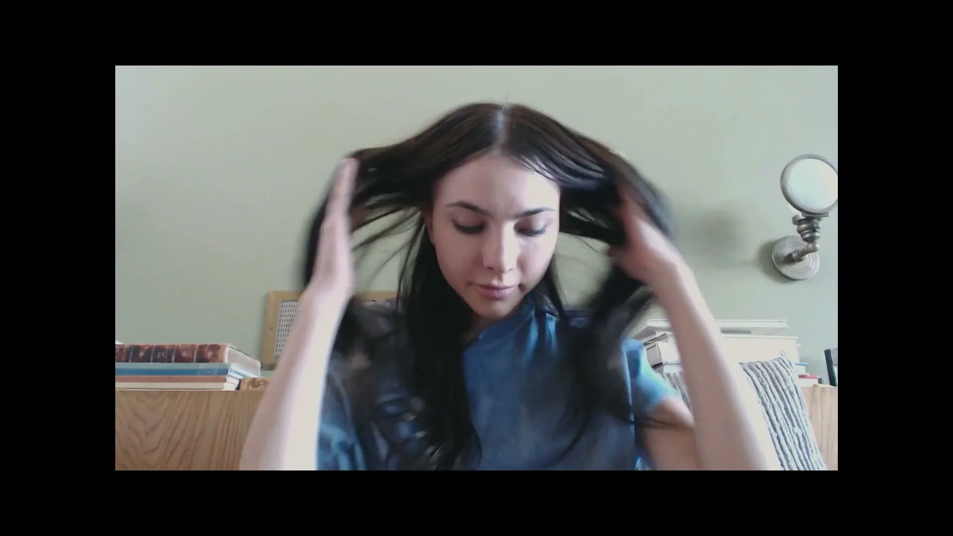 A young woman with long dark hair, wearing a blue shirt, looks into the camera and runs her hands through her hair in a bedroom.