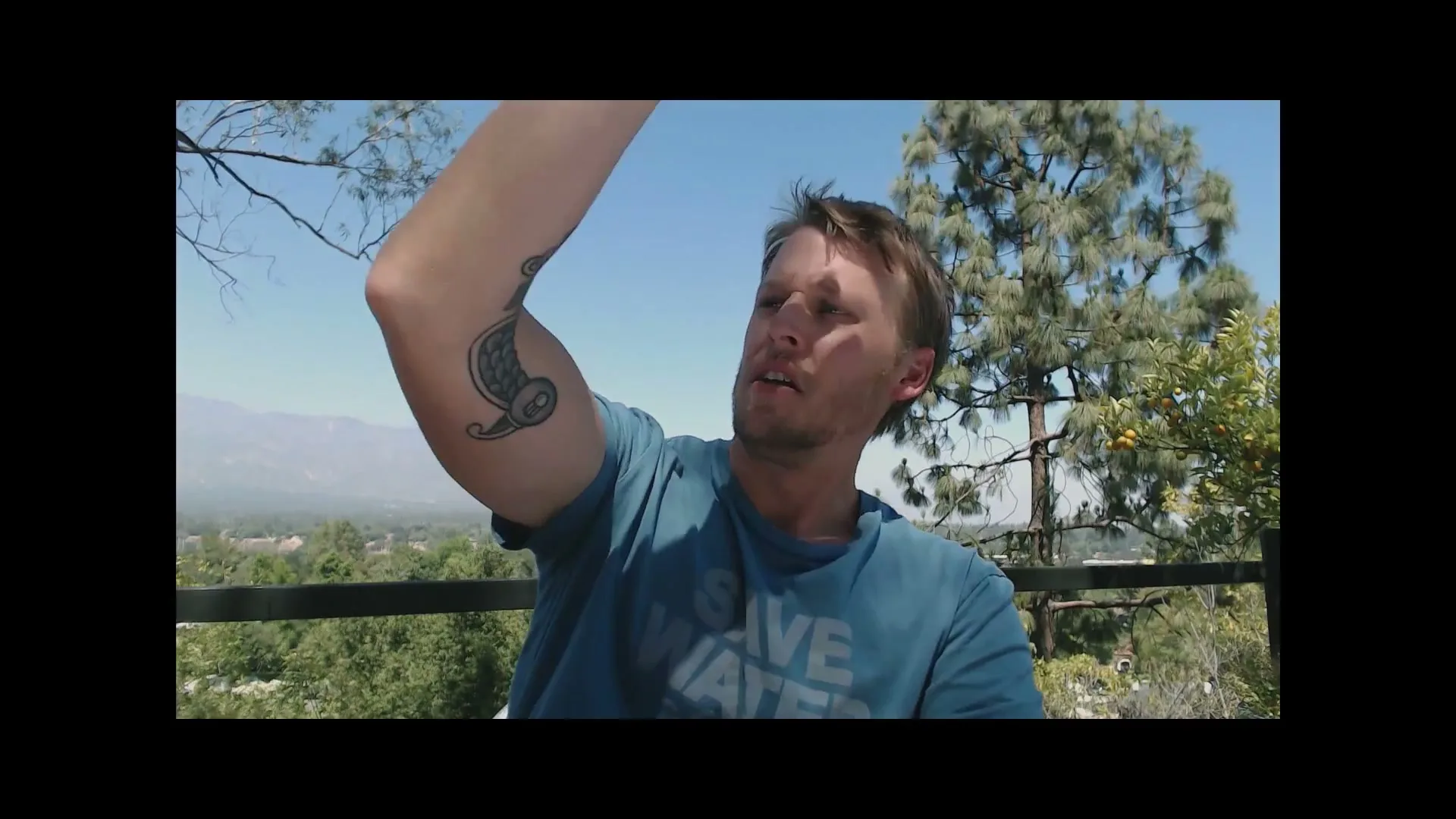 A man with a beard and a blue t-shirt with a fish tattoo on his arm looks up and shields his eyes from the sun on a balcony overlooking a valley.