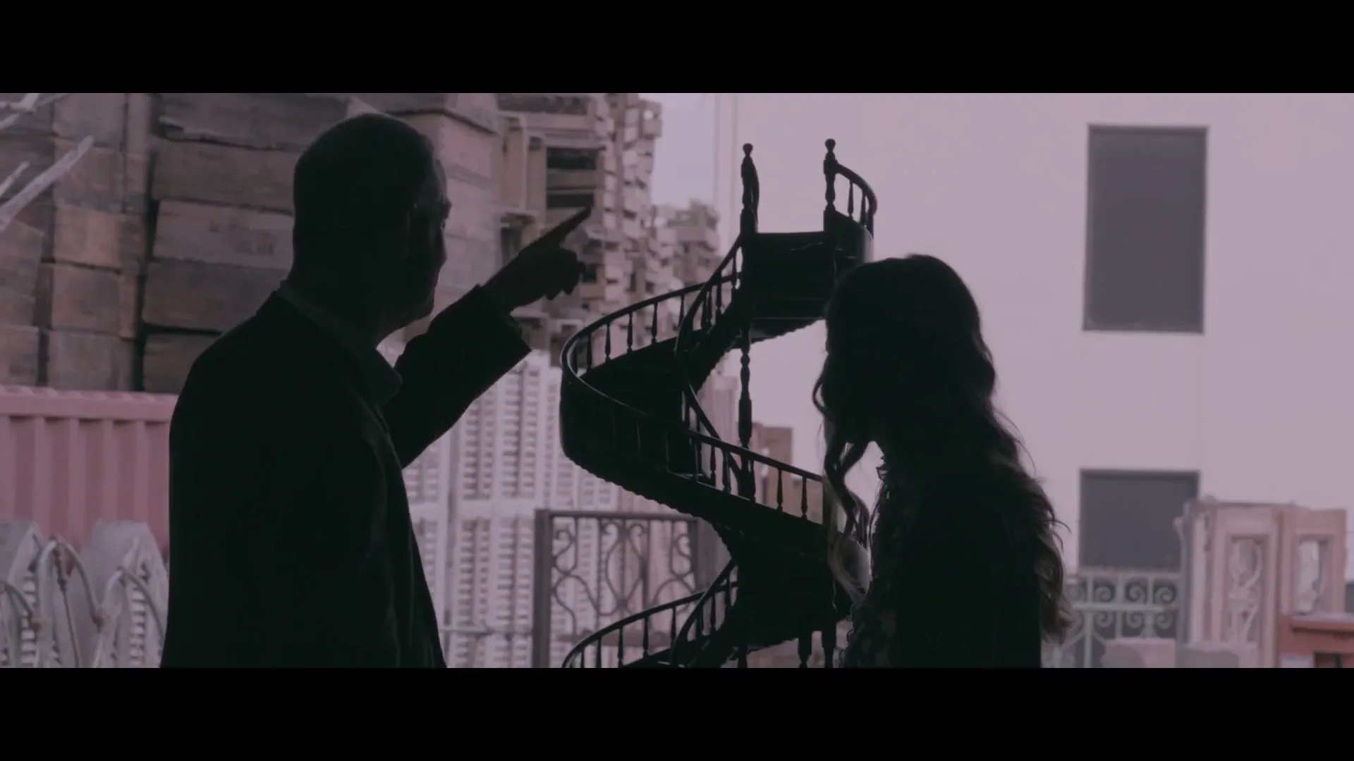A man and a woman are silhouetted against a bright background, looking at a spiral staircase in an outdoor salvage yard.