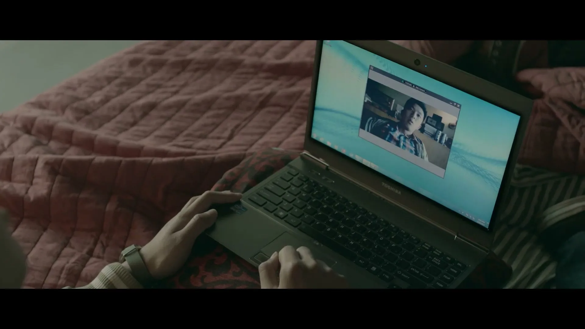 A person's hands are seen typing on a Toshiba laptop, which displays a video call with a young man on the screen, while resting on a red patterned blanket.