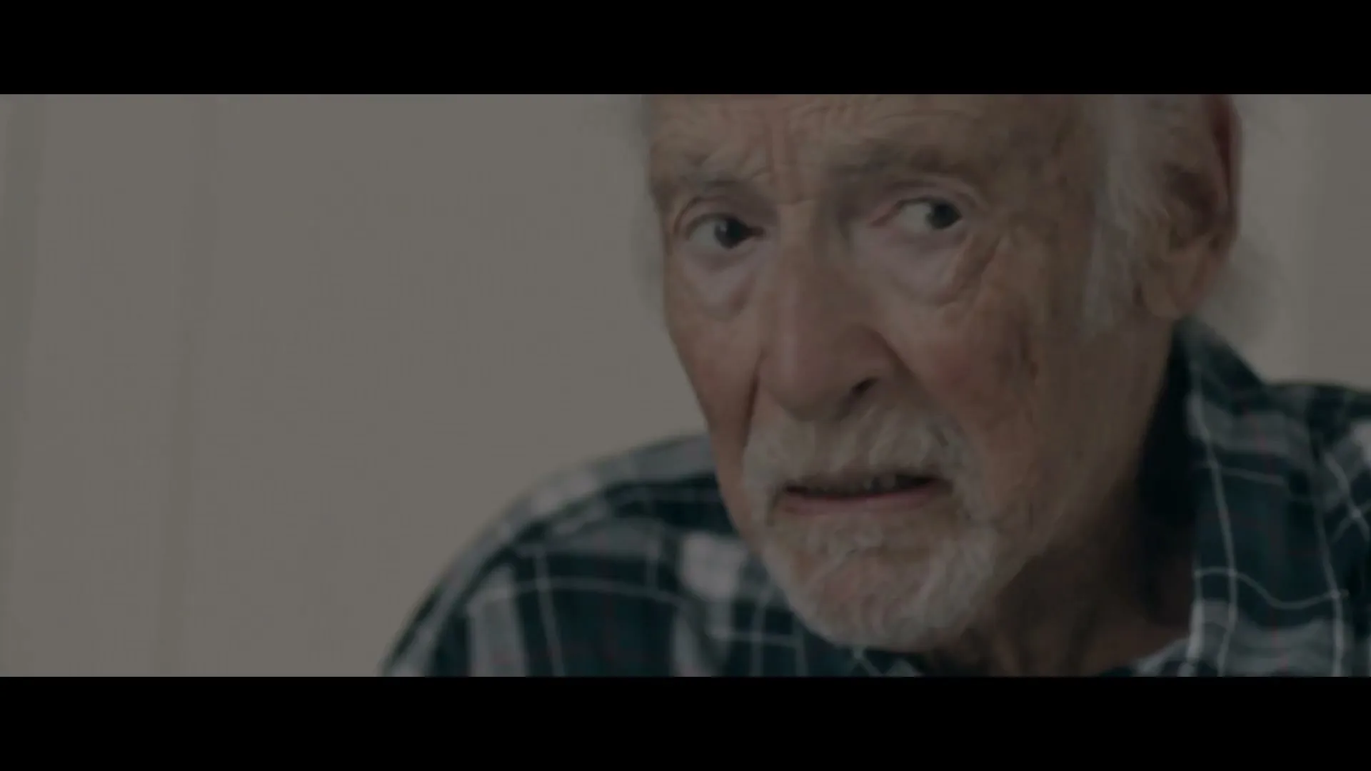 An elderly man with white hair and a beard, wearing a plaid shirt, looks off-camera with a concerned expression in an interior setting.