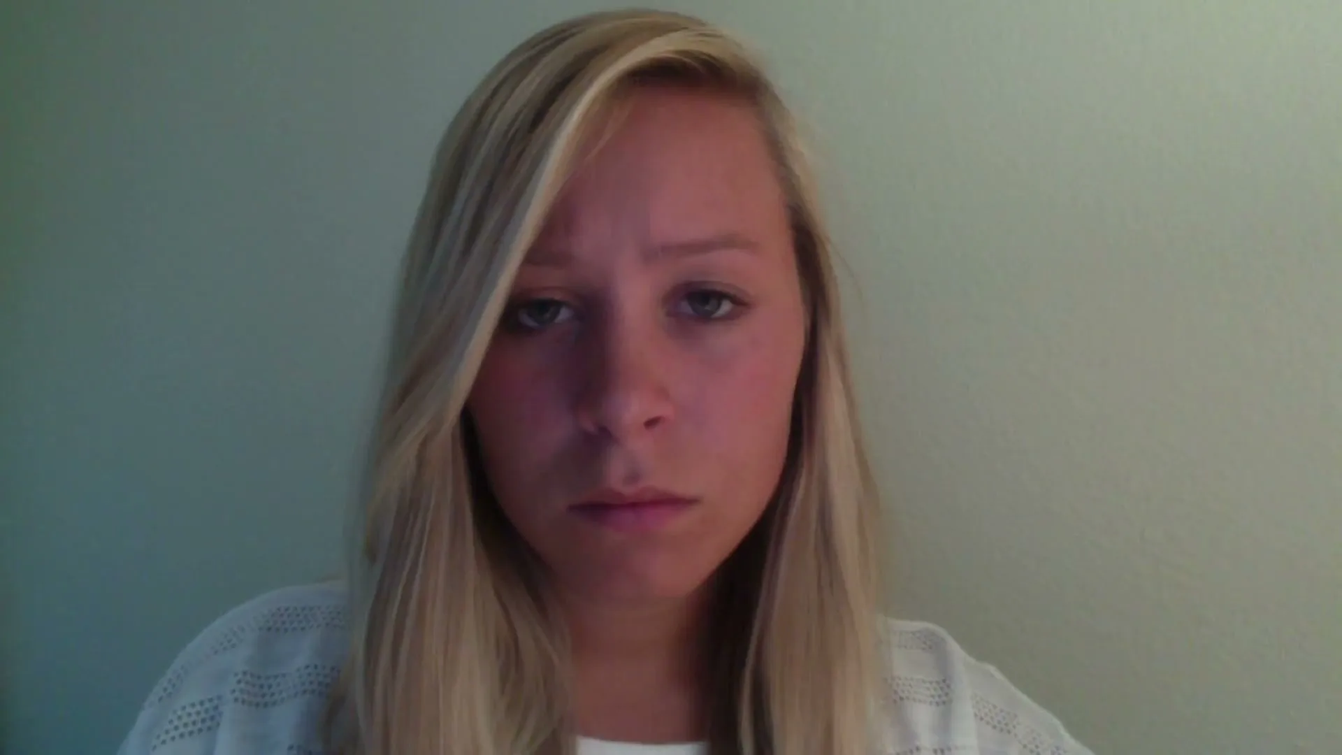 A young blonde woman with a serious expression looks directly at the camera, wearing a white top in a simple interior setting.