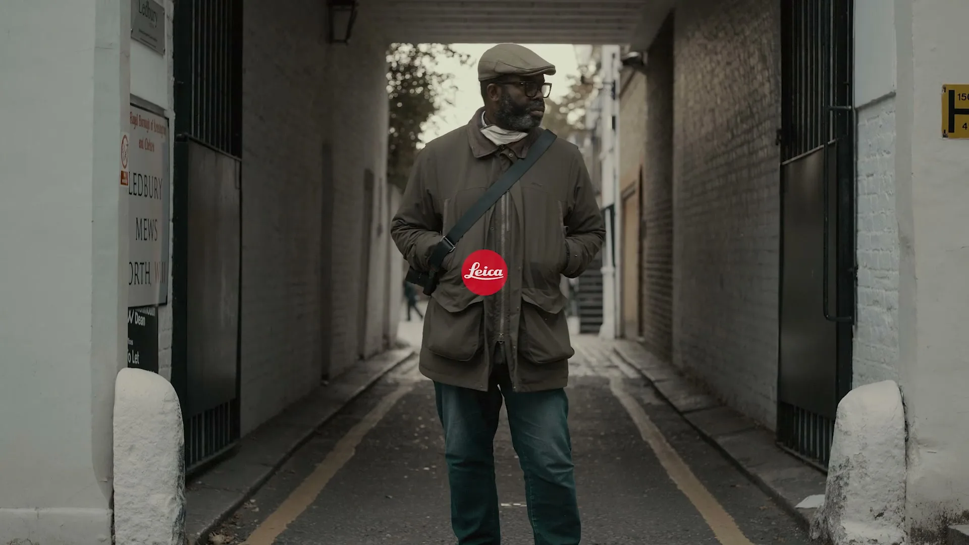 A man with a beard and glasses stands in a narrow alleyway, wearing a coat and a cap, looking to the right.