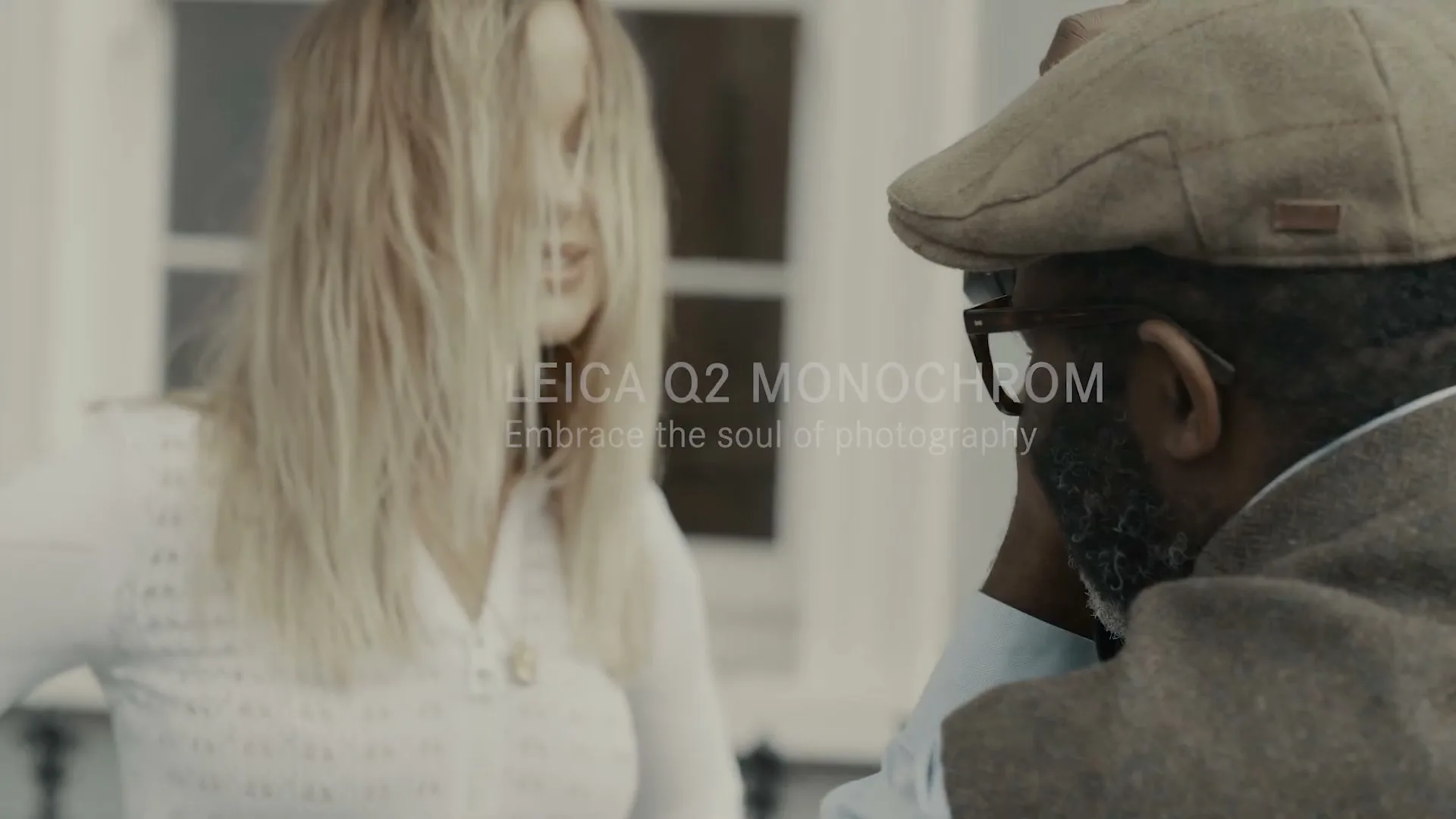 A blonde woman with hair covering her face talks to a man in a flat cap and glasses, both in soft focus, with text overlay.