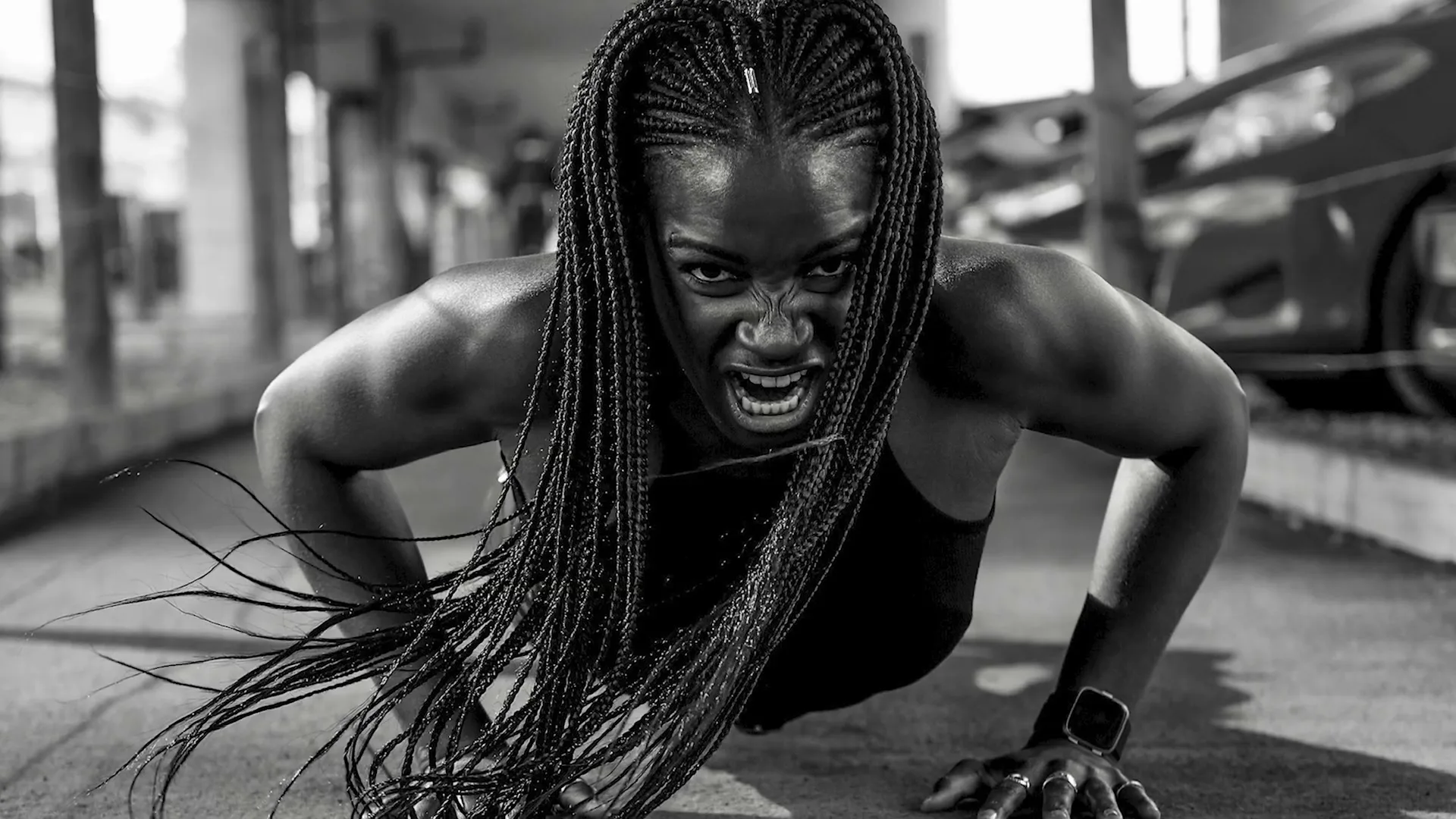 A determined Black woman with braided hair, wearing a tank top, does push-ups on the ground in an urban setting, looking intensely at the camera.