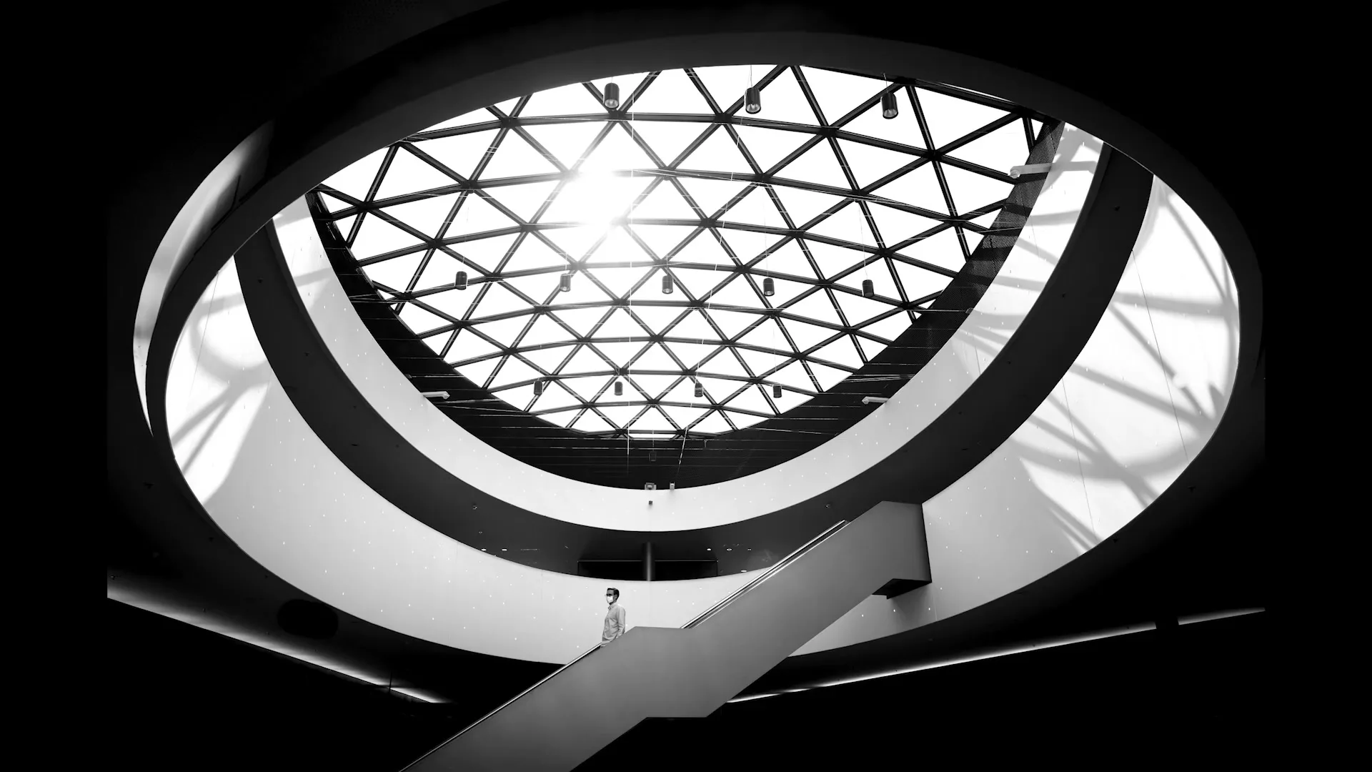 A lone person stands on an escalator in a modern, circular building with a geometric glass ceiling, captured in black and white.