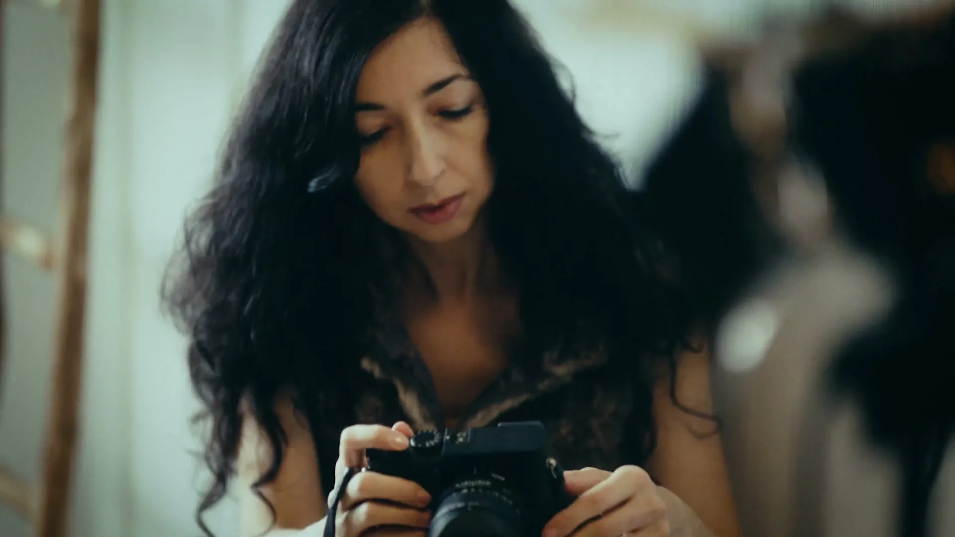 A woman with long dark hair, wearing a fur vest, looks down intently at a black camera she holds in her hands, in an interior setting.