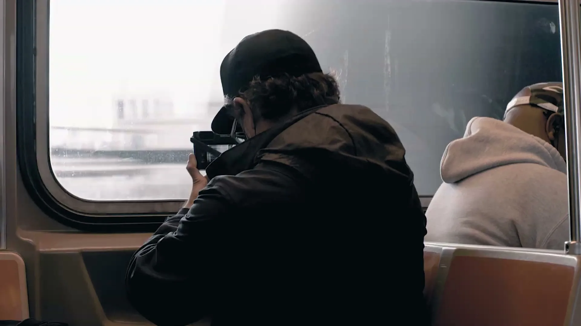 A man in a black cap and jacket sits on a train, holding a camera and looking out the window as the train moves.