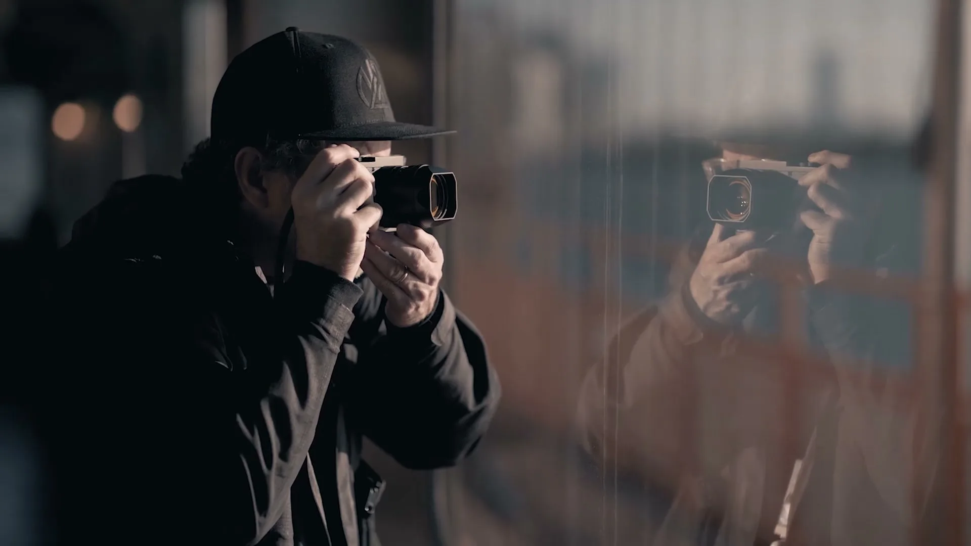 A man in a black cap and jacket holds a camera to his eye, reflected in a window, taking a photo.