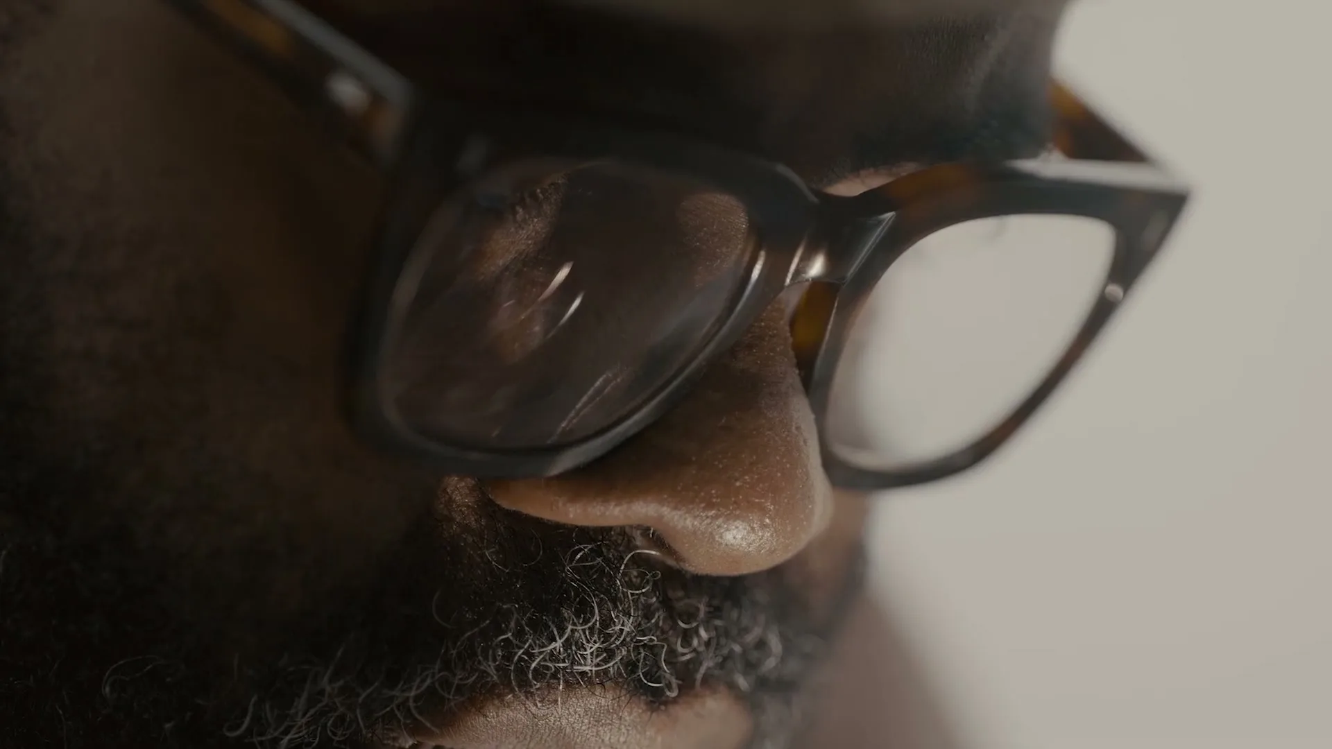 A man with a beard and glasses is shown in a close-up shot, looking down, in an interior setting.