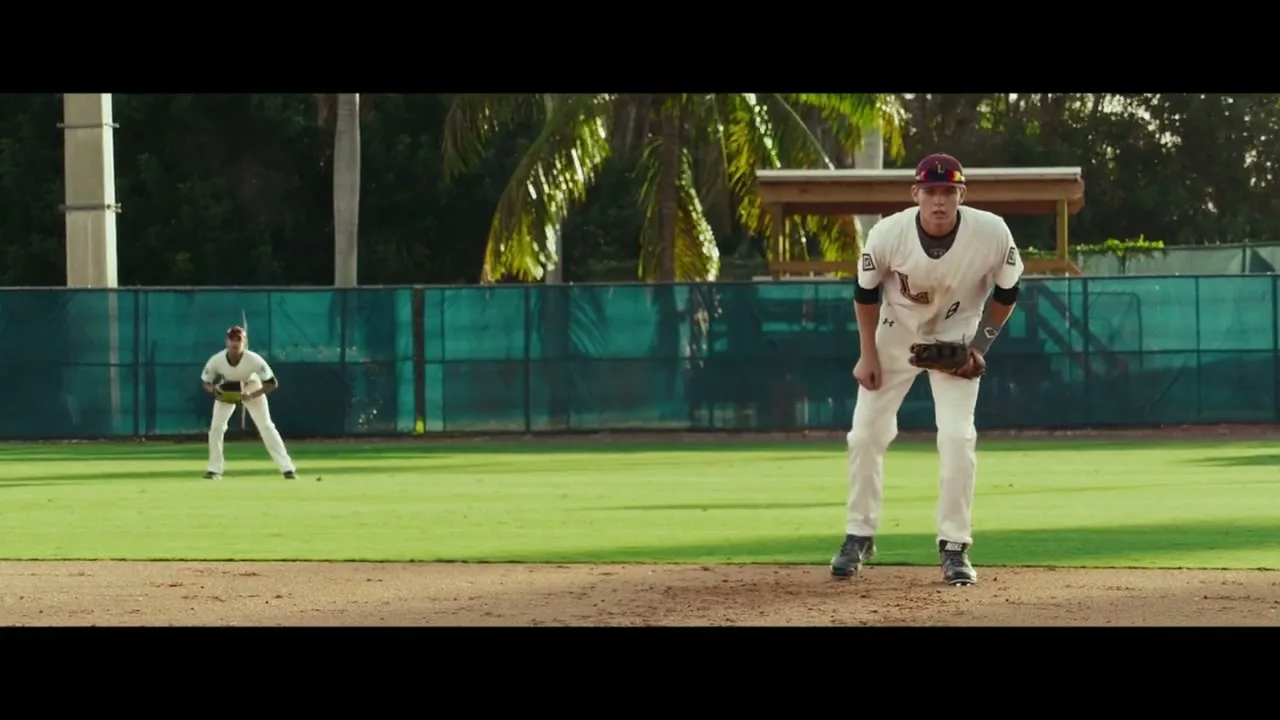 A male baseball player in a white uniform and cap stands on the pitcher's mound, looking focused, while another player is in the background on a sunny baseball field.