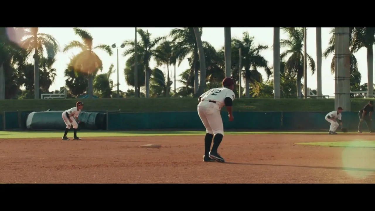 A baseball player in a white uniform with the number 2 on his back stands on a dirt field, with other players and palm trees in the background.
