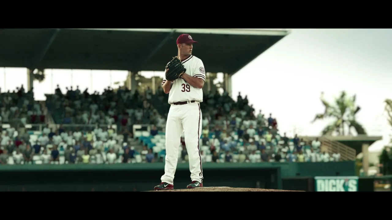 A male baseball pitcher in a white uniform with a maroon cap and glove stands on the mound, preparing to pitch in a stadium full of spectators.