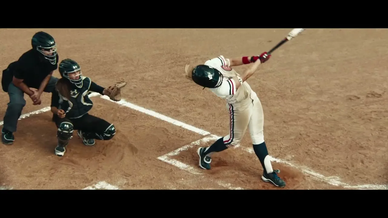 A female softball player in a white and blue uniform swings a bat, hitting a yellow softball, while a catcher and umpire watch on a dirt field.