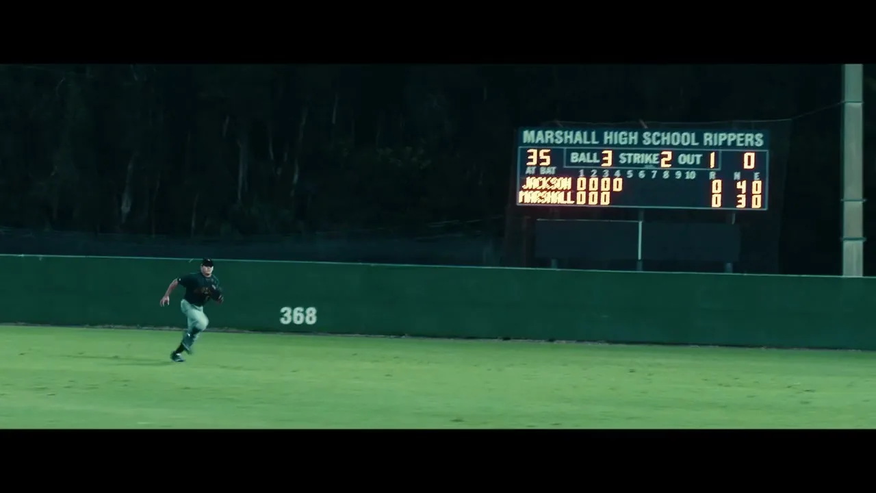 A male baseball player in a dark uniform runs across a green field at night, with a scoreboard visible in the background.