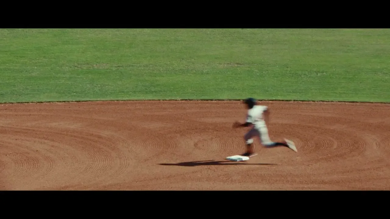 A blurry male baseball player in a white uniform runs past a white base on a dirt field under a bright sky.