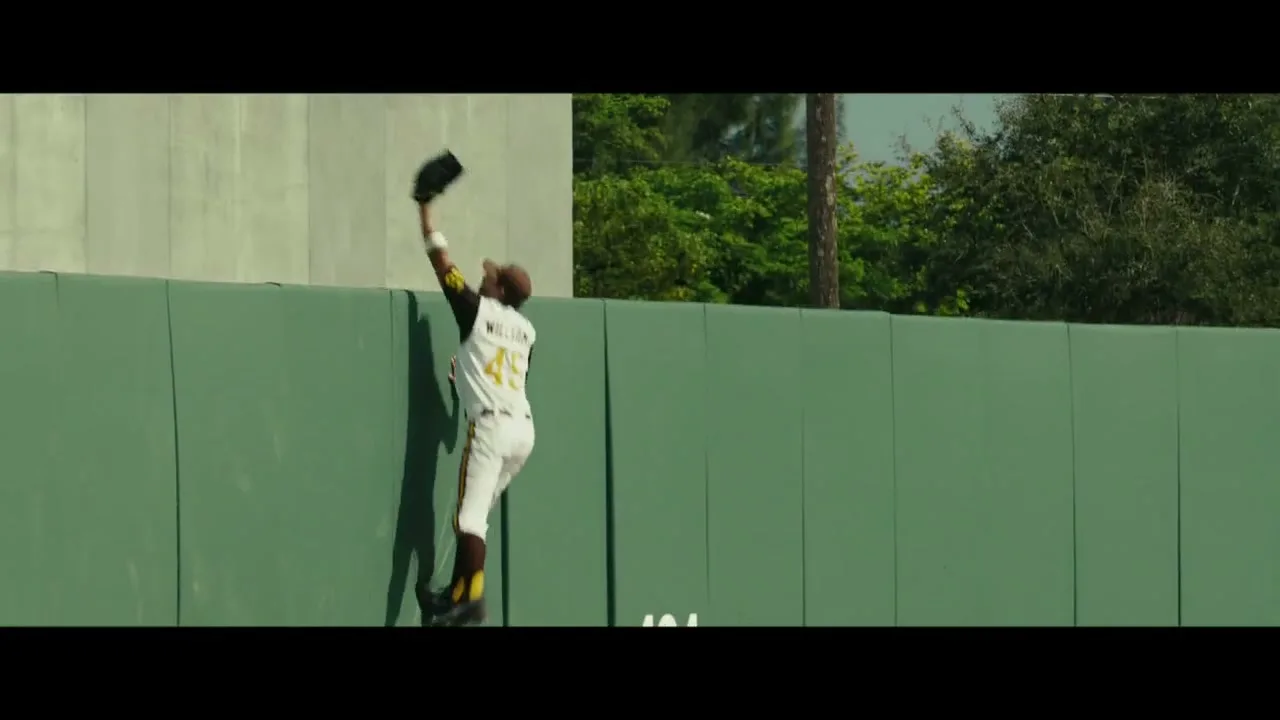 A male baseball player in a white and yellow uniform with number 45 jumps against a green outfield wall to catch a ball during a game.