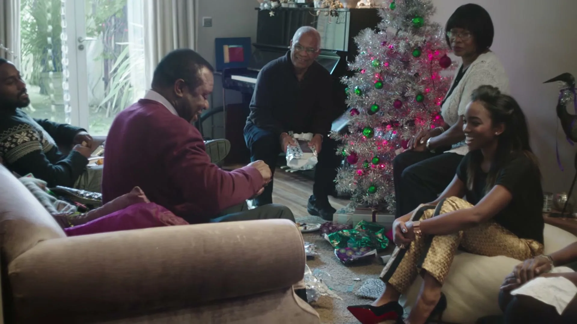 A multi-generational Black family gathers in a living room, exchanging gifts around a silver Christmas tree, with one man unwrapping a present.