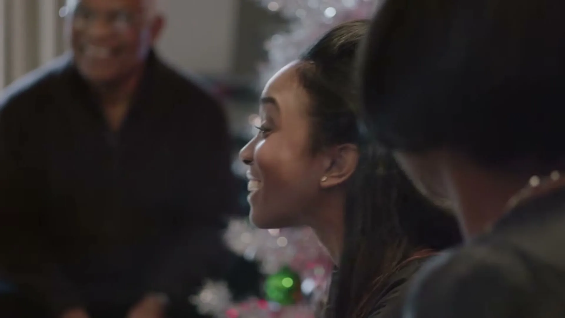 A young Black woman with long dark hair smiles and looks to the right, while an older Black man smiles in the blurred background, next to a white Christmas tree.