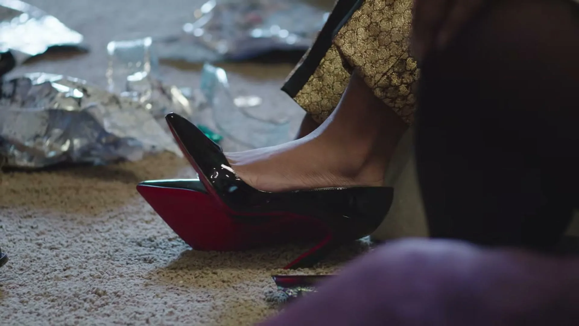 A woman's foot, wearing a black high heel with a red sole, is seen on a beige carpet amidst discarded wrapping paper and gift boxes.