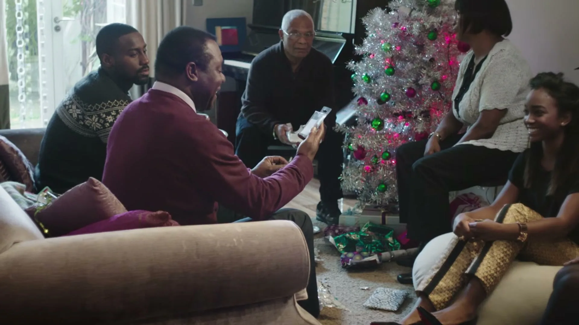 A family of five, dressed in casual attire, sits around a silver Christmas tree in a living room, exchanging gifts.