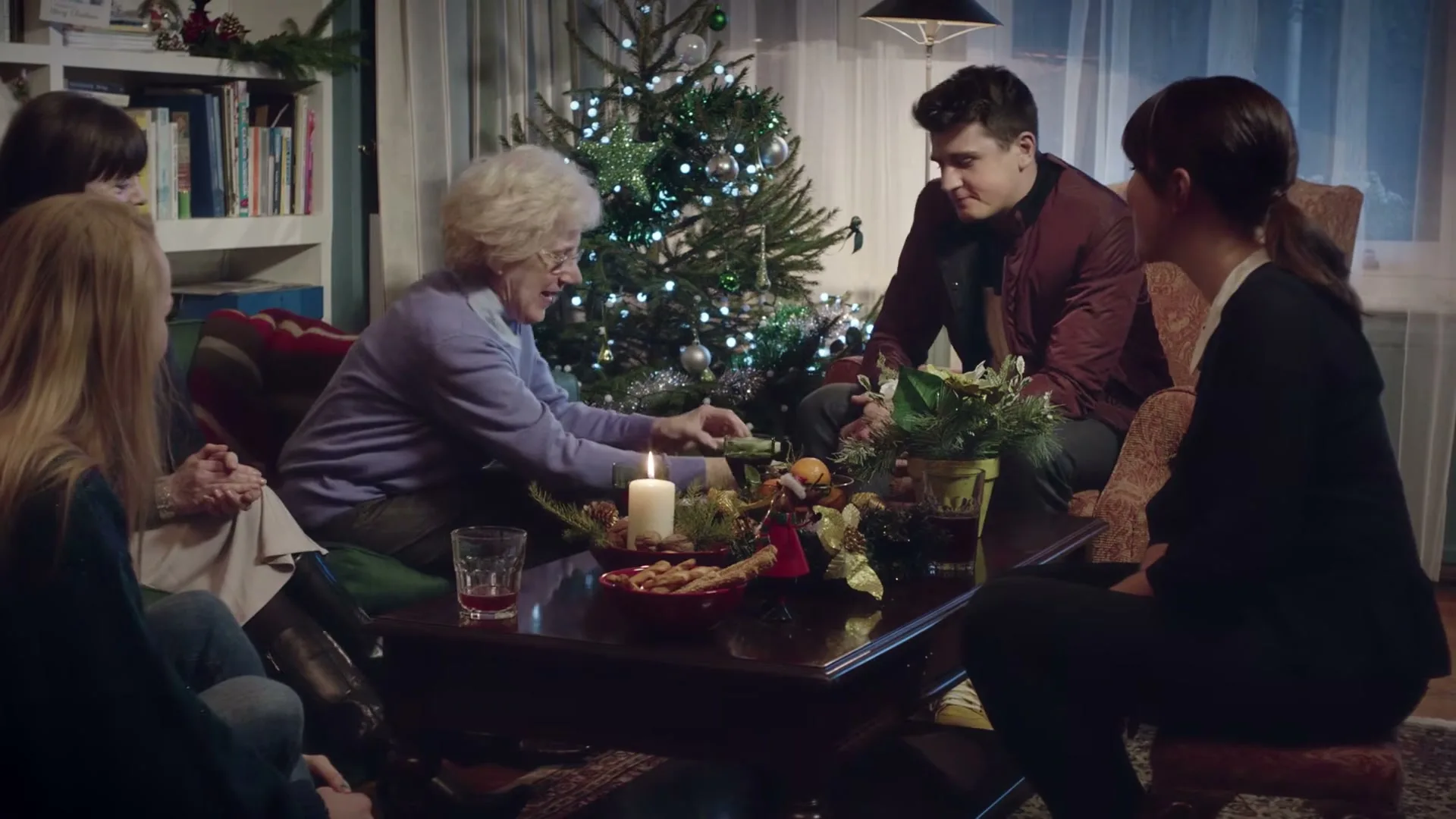 An elderly woman in a purple sweater opens a gift while sitting on a couch with three other people around a coffee table with a lit candle and snacks, next to a decorated Christmas tree.