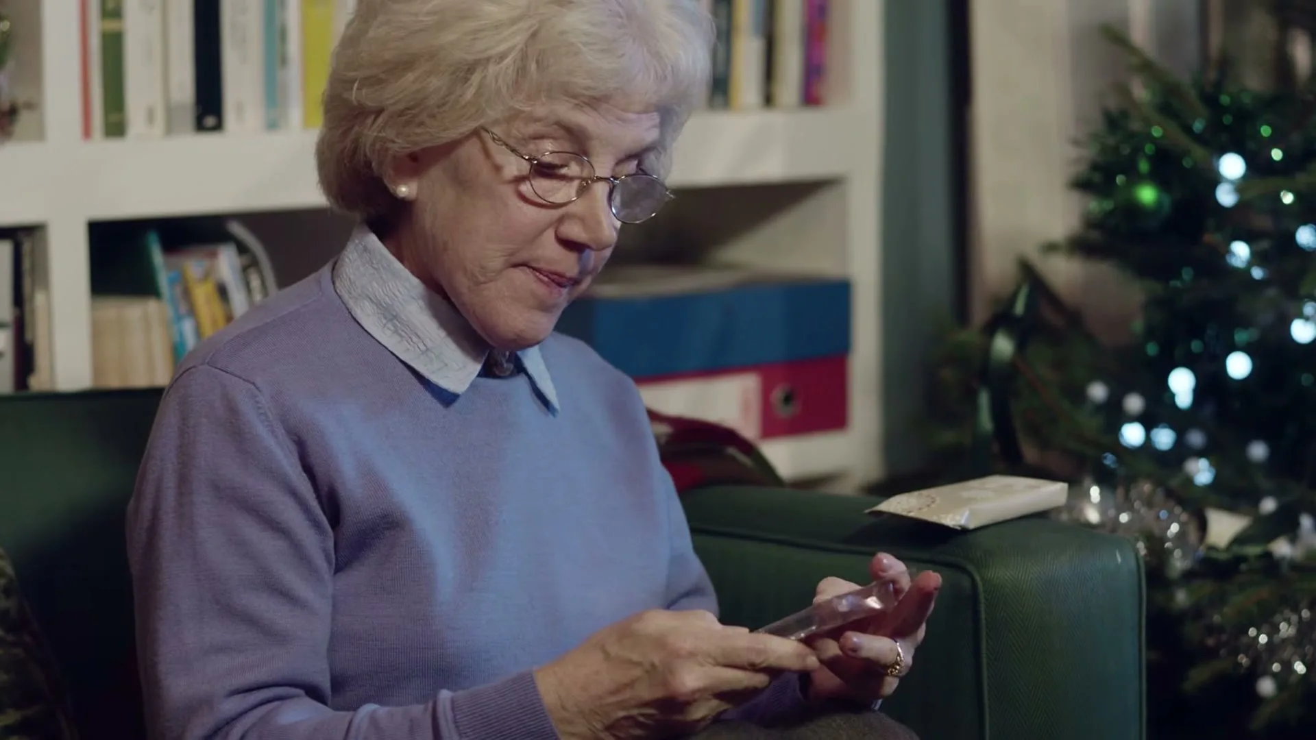 An elderly woman with white hair and glasses sits on a green couch, opening a small, clear plastic package of paperclips, with a Christmas tree in the background.