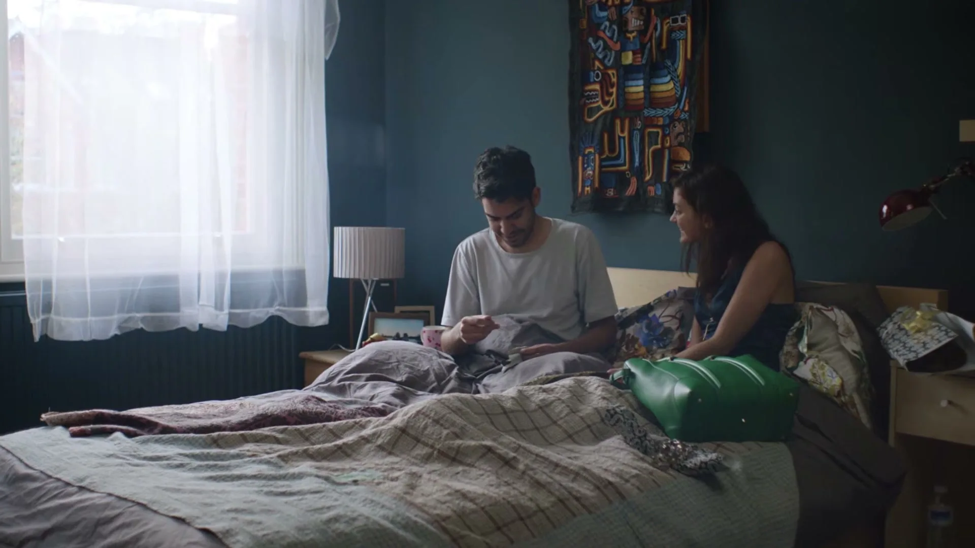 A man and woman sit on a bed in a bedroom, the man looking at something in his hands while the woman smiles at him.