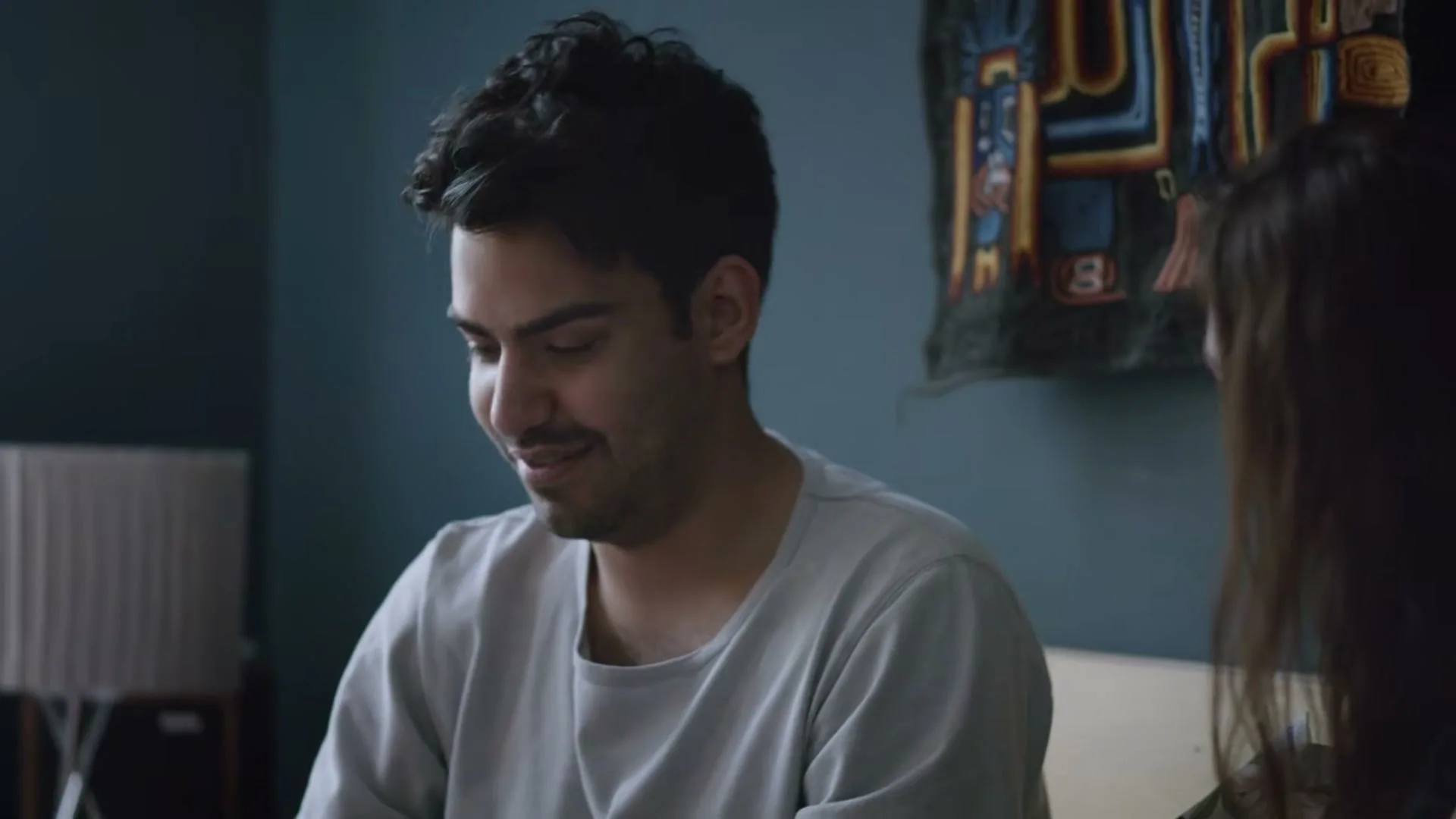 A young man in a grey t-shirt sits on a bed, looking down at a small box, then smiles at a woman next to him.