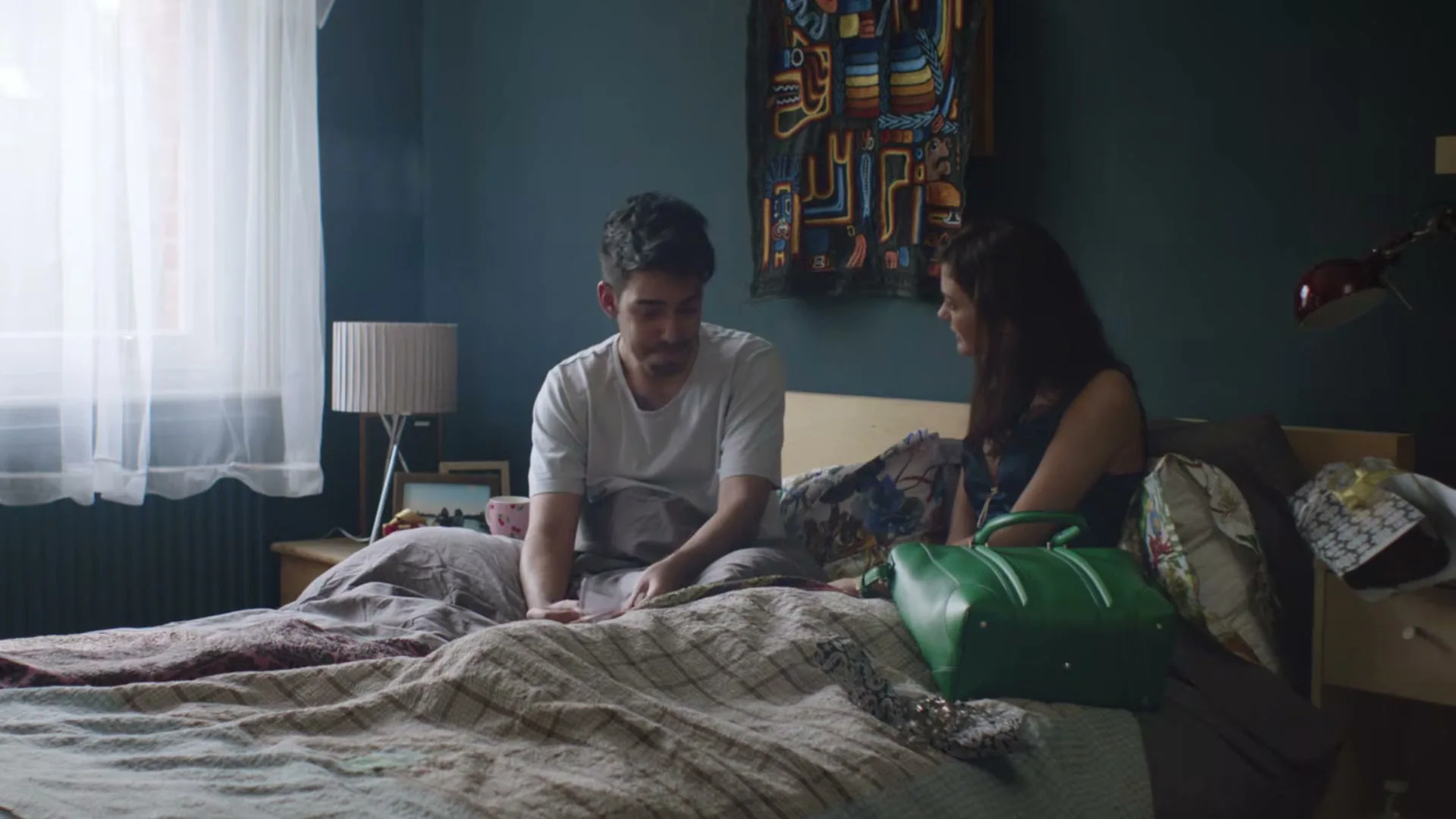 A man in a white t-shirt sits on a bed, looking down, while a woman in a dark dress talks to him, next to a green handbag in a bedroom.