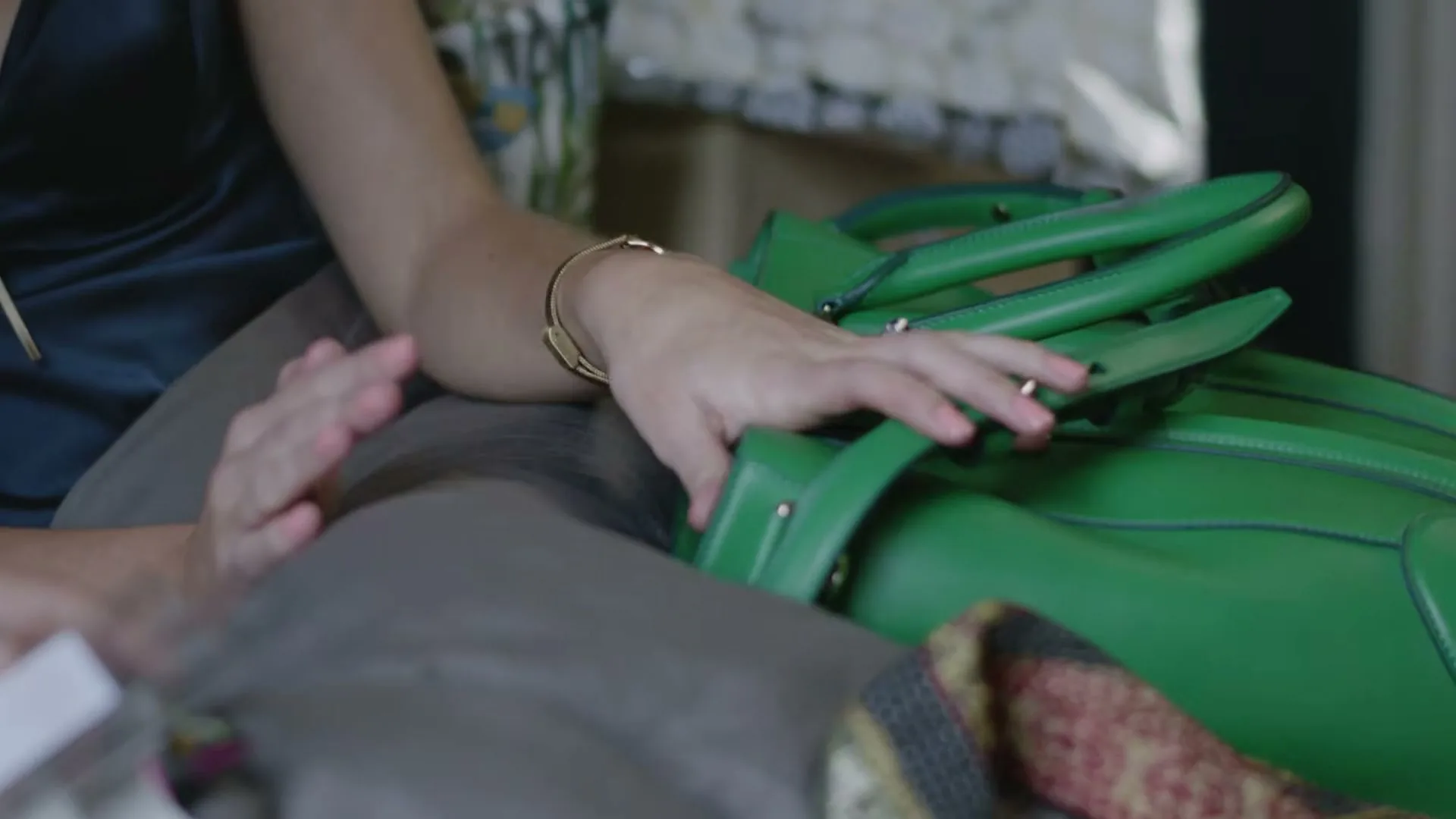 A woman in a dark blue top and brown pants sits, admiring a bright green handbag on her lap, in a cozy interior setting.