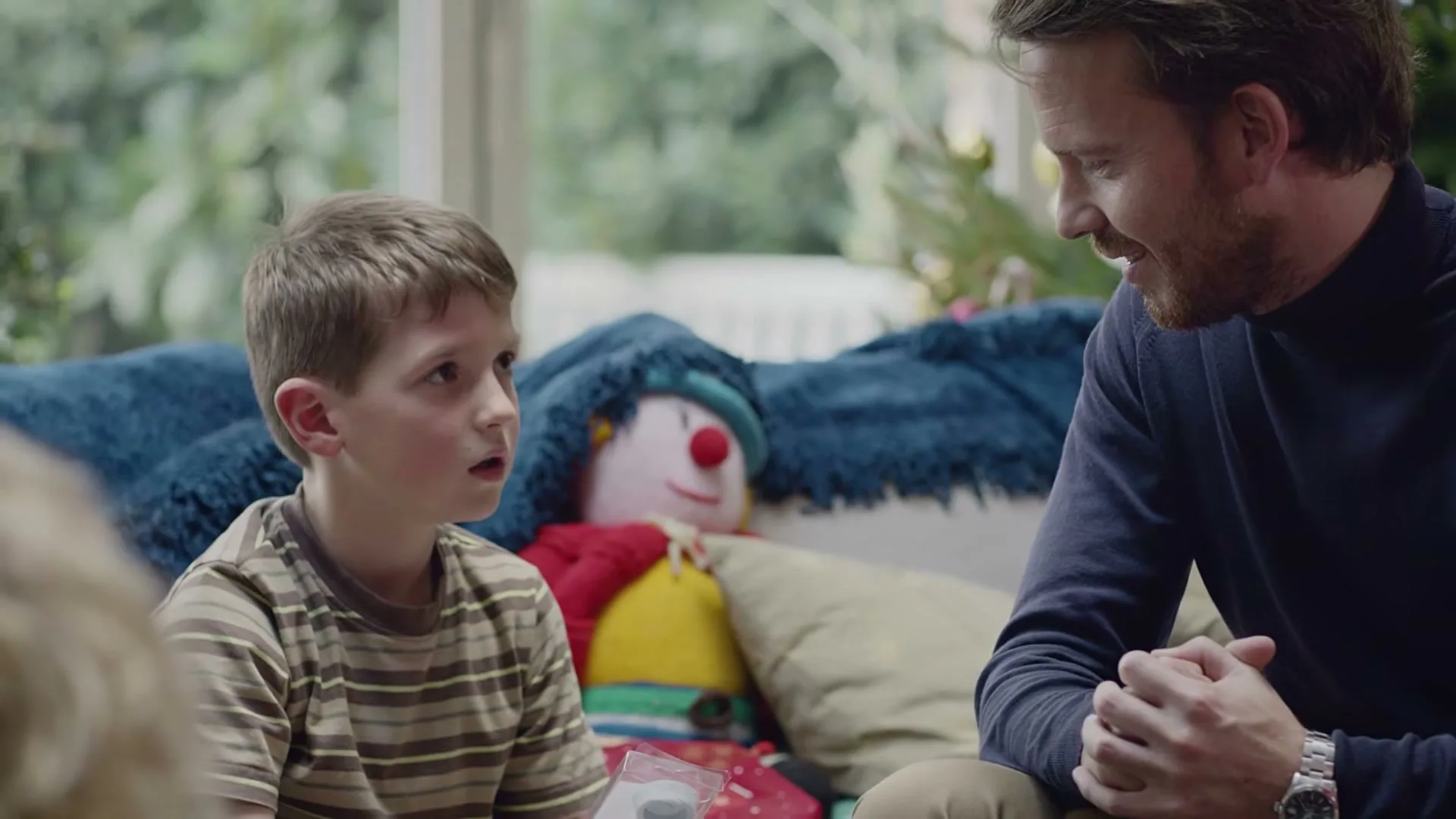 A young boy with a striped shirt looks confused while opening a small gift next to a smiling man in a living room.