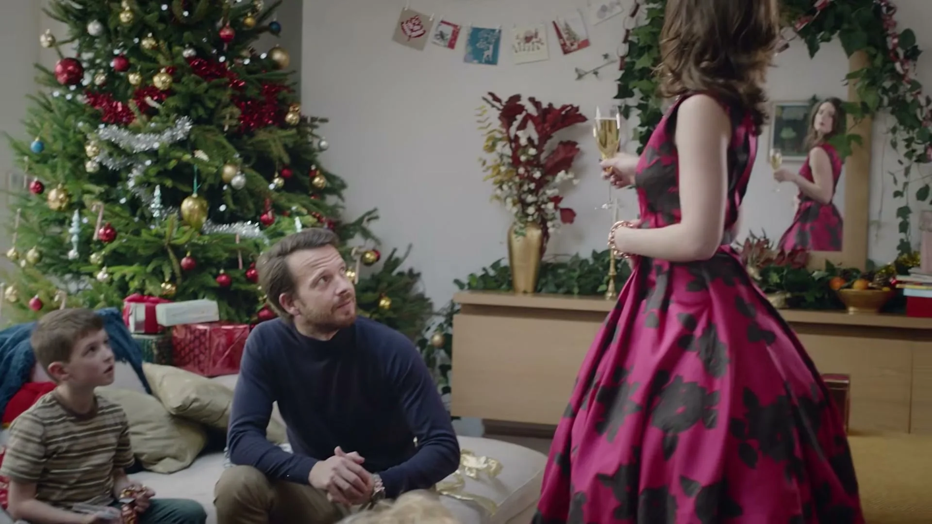 A woman in a pink and black dress stands in a living room, holding a champagne glass, while a man and a boy sit on a couch, looking at her.