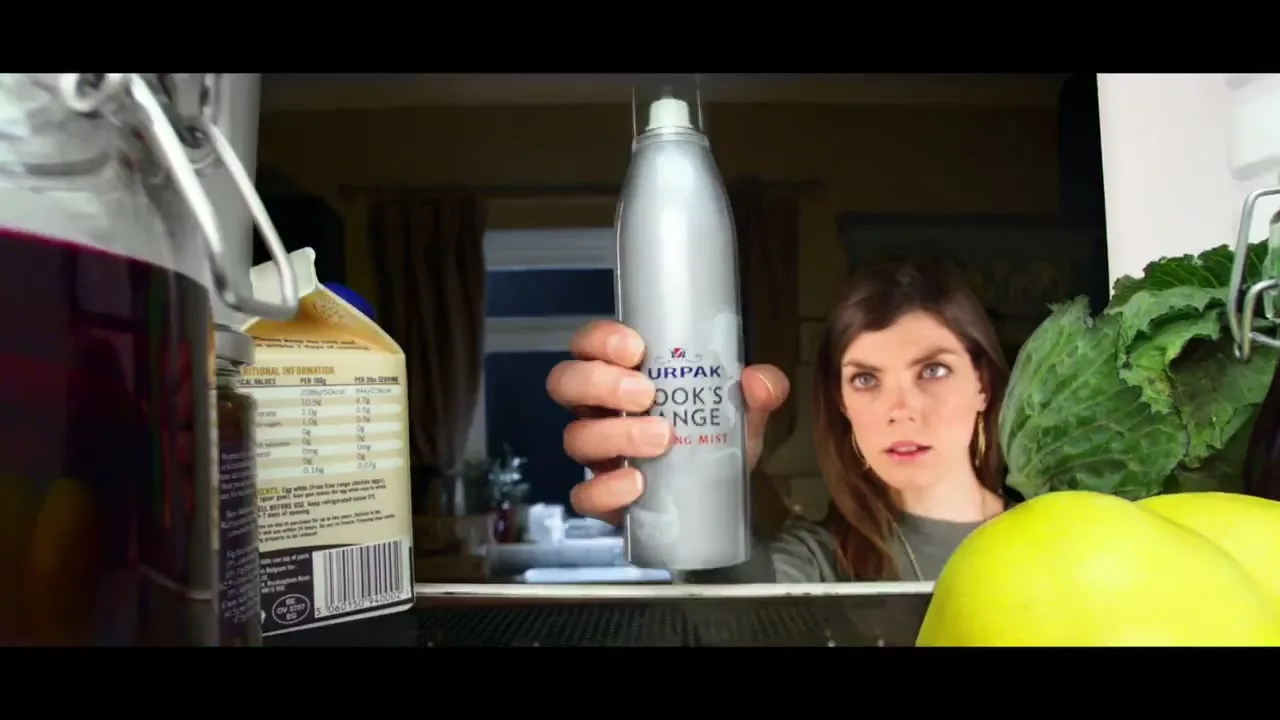 A woman with brown hair and a green shirt looks surprised as she holds a silver spray bottle of Lurpak Cook's Range Cooking Mist inside a refrigerator.