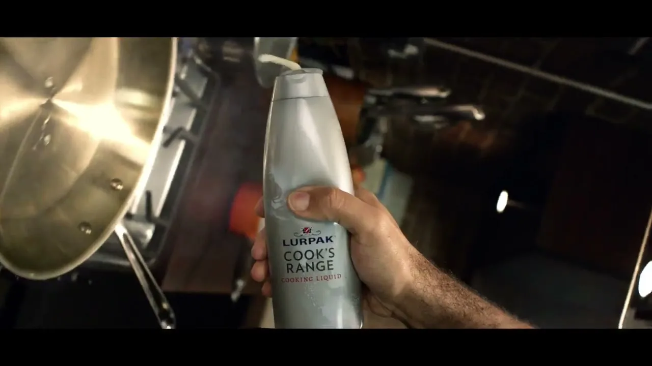 A man's hand holds a bottle of Lurpak Cook's Range cooking liquid, squeezing it into a pan on a stovetop in a kitchen.
