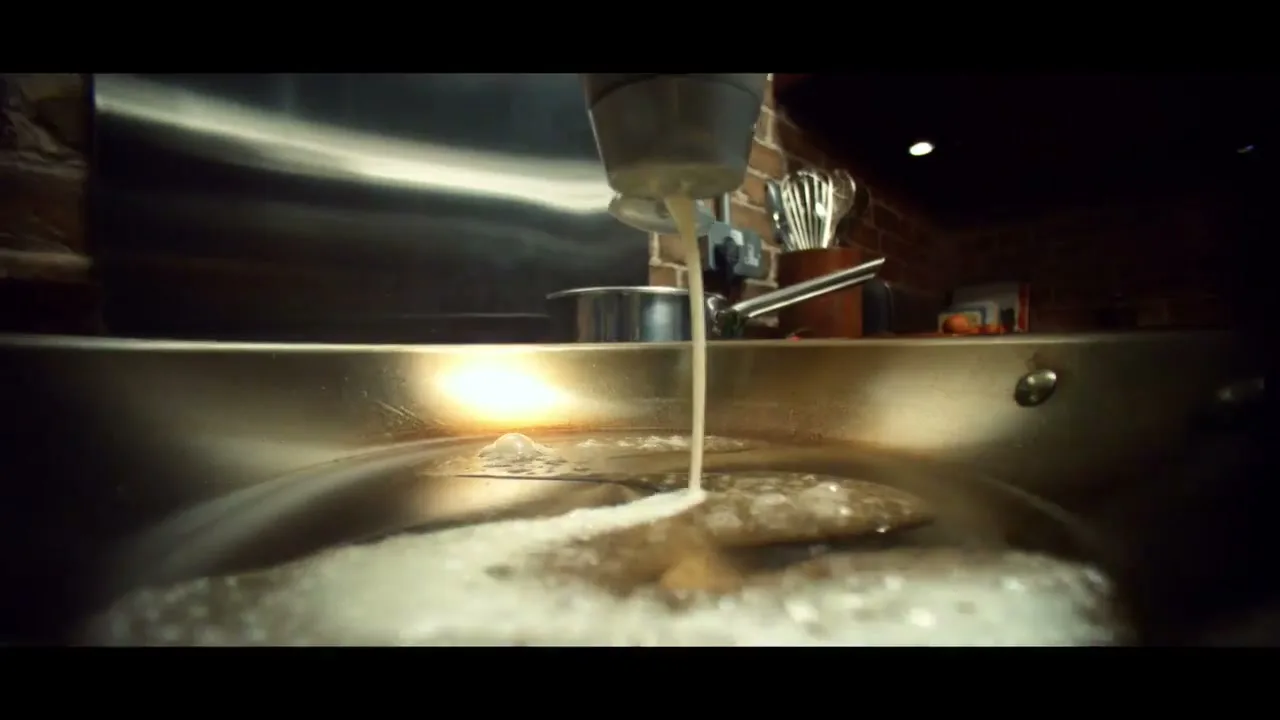 A close-up shot shows a white liquid being poured from a metal container into a sizzling pan, creating bubbles and foam.