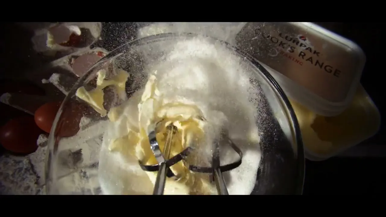 An overhead shot shows an electric mixer beating butter and sugar in a glass bowl, with eggs and a Lurpak butter tub nearby.